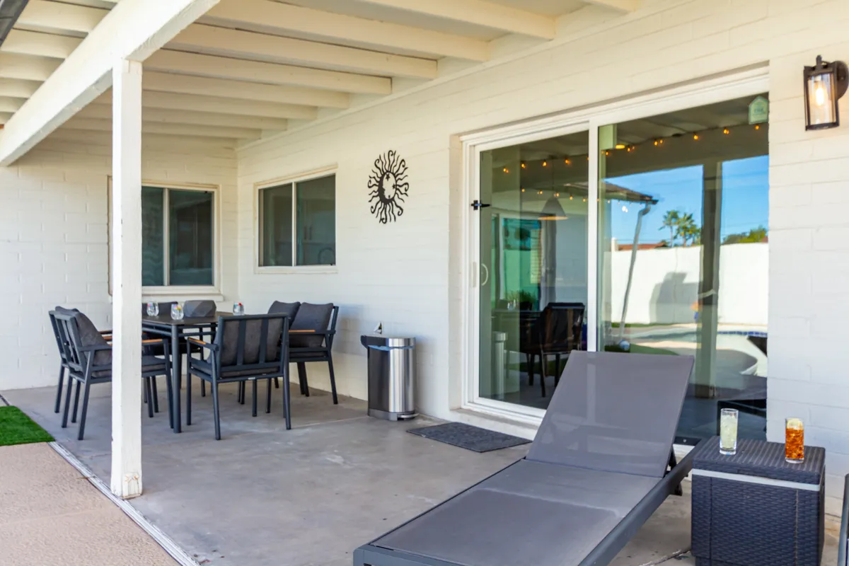 A covered patio with a modern dining table and chairs sits adjacent to a lounge chair and a sliding glass door leading back inside. The patio is decorated with a wall clock and offers ample seating for alfresco dining. The relaxed setup invites guests to enjoy meals outdoors in comfort.