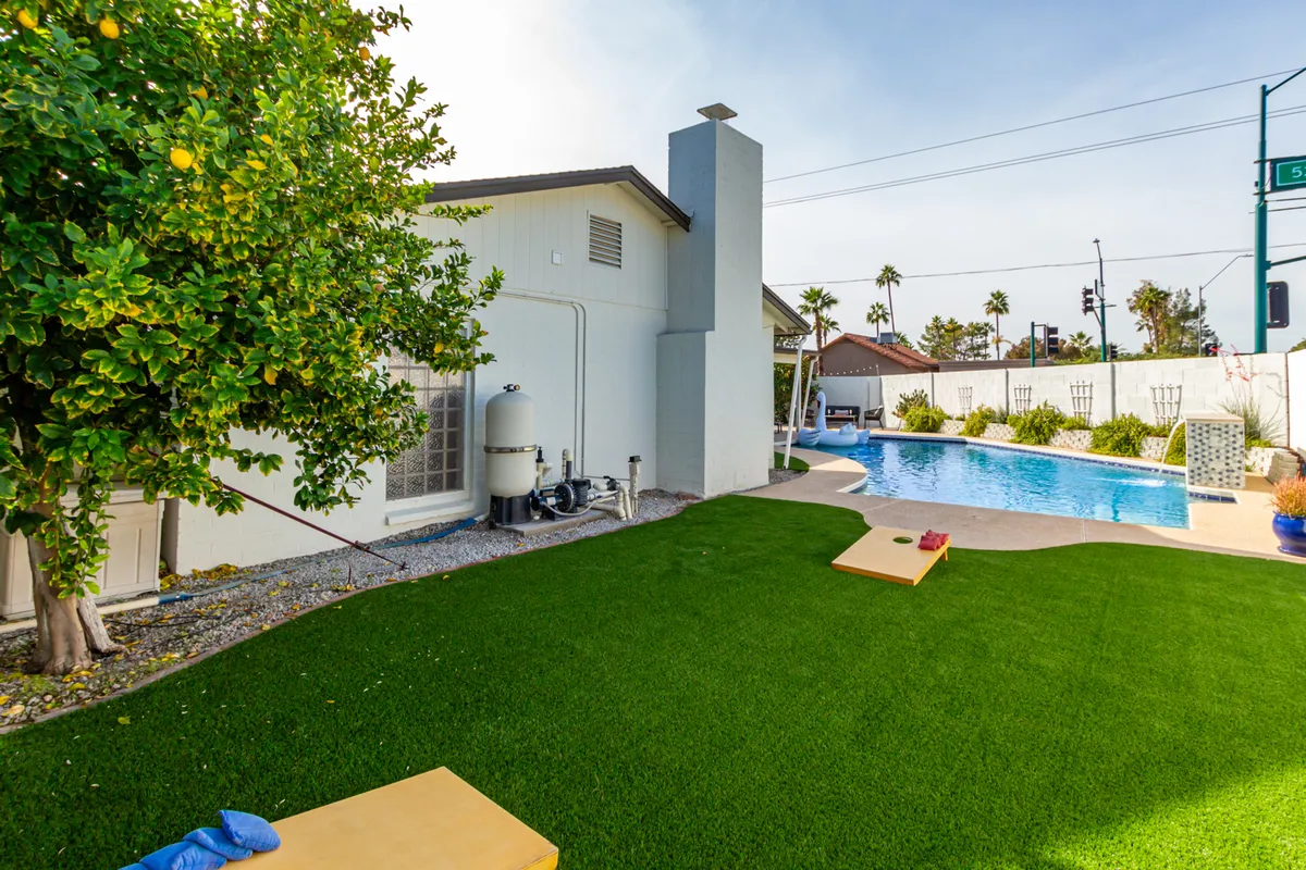 A spacious backyard scene features a sparkling swimming pool bordered by lush greenery and a manicured lawn. The lawn area is set up for outdoor games like cornhole, and there is a stunning view of the pool with the house's chimney visible in the background. The sun is low, casting a warm, inviting glow that sets the tone for both daytime fun and evening relaxation.