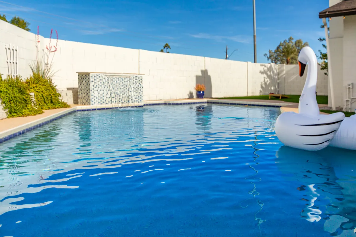 A vibrant view of the backyard swimming pool, accented by a playful swan inflatable and a tranquil waterfall feature. Lush greenery surrounds the pool area, creating a sense of privacy and resort-like ambiance. The scene captures both the playful and relaxing elements of the backyard.