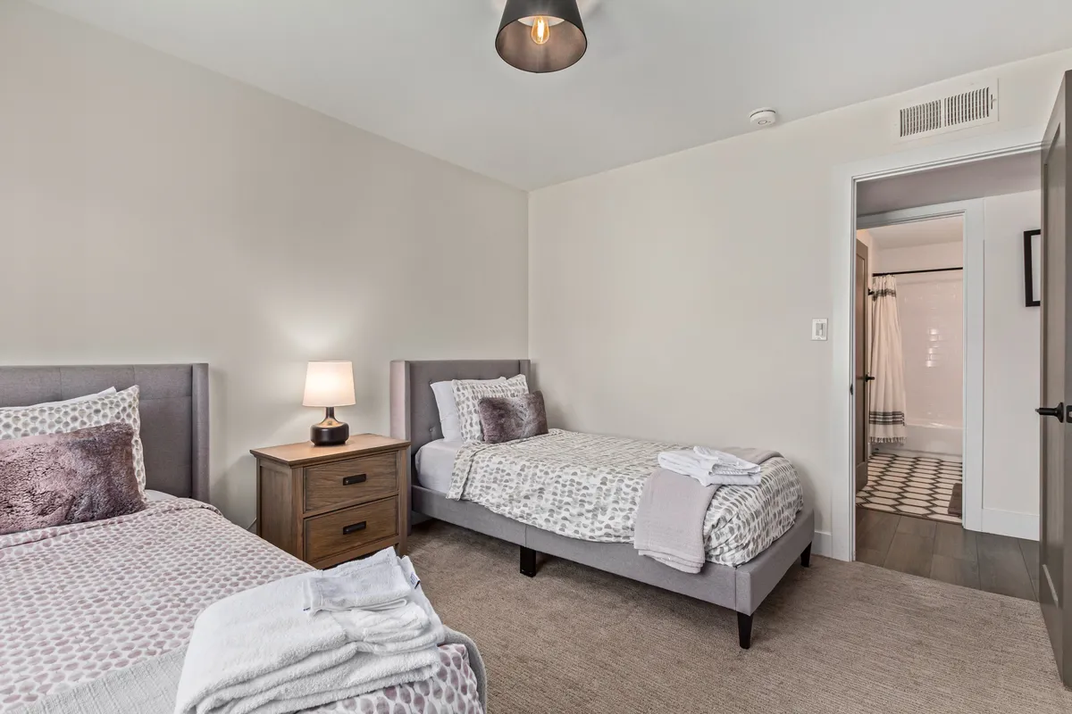 A cozy bedroom with two twin beds, a shared nightstand with a lamp, and a visible bathroom doorway, giving the space a welcoming and versatile appeal. The natural light and neat arrangement suggest suitability for both families and friend groups.