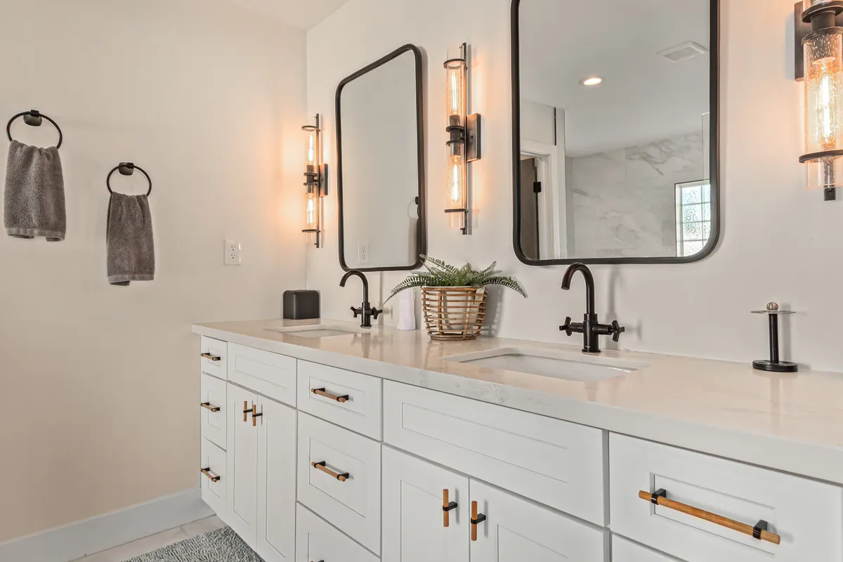 A spacious, modern bathroom with double sinks, large mirrors, stylish light fixtures, and plush towels. Decorated with a touch of greenery, the space feels fresh, bright, and inviting—ideal for morning and evening routines.